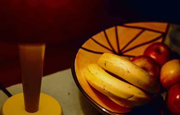 Eine Hotelatmosphaere: Die Tischlampe mit rotem Lampenschirm verschmilzt mit ihrem Hintergrund in dunklem Rot. Neben der Lampe steht eine gelborangene Obstschale, gefuellt mit Bananen im Vordergrund und Aepfeln im Hintergrund, unscharf. Die Obstschale ziert eine spinnennetzartige Ornamentik. Die Tischlampe hat einen gelben Fuss