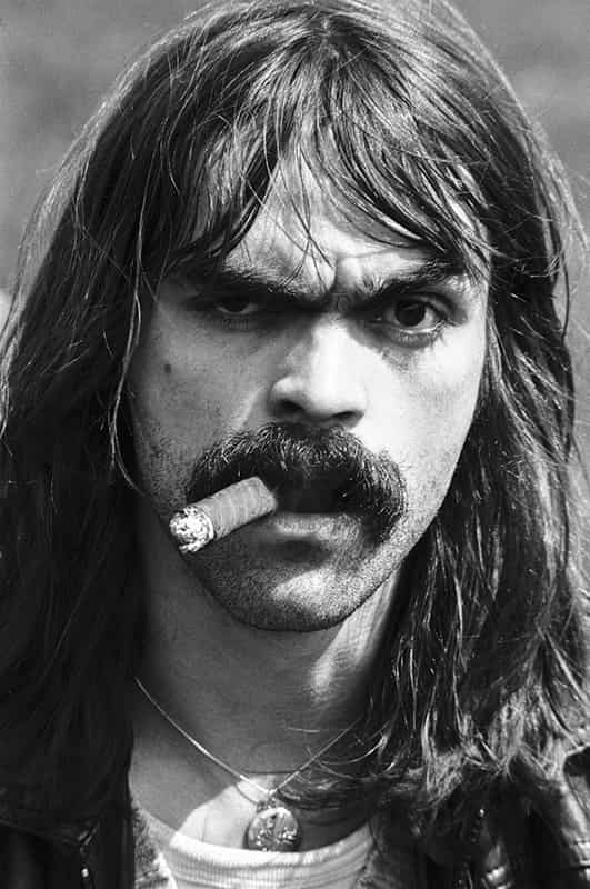 Sein Nickname ist Diddi. Seinen ovalen Kopf umsaeumen lange schwarze Haare, er traegt einen Mittelscheitel, Haare fallen ihm links und rechts auf die Stirn. Das linke Auge ist weit geoeffnet, das rechte Auge zusammen gekniffen. Die linke Augenbraue ist hochgezogen - beide sind schwarz. Von rechts einstroemendes Licht moduliert sein Gesicht. Ein gewaltiger, dichter schwarzer Schnurbart kroent seine Oberlippe und umrahmt die Unterlippe. In seinem Mund steckt eine Zigarre - Monte Christo, Numero 2 Special - der Rauch verdeckt seine Zaehne. Ein Dreitagesbart perfektioniert das Erscheinungsbild. Um den Hals traegt Diddi ein Amulett.