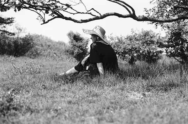 Eine unbefahrene Strassenkreuzung auf Menorca, in der Mitte befinden sich zwei nebeneinander liegende Gullydeckel. Ein zweigeschossiges Haus ohne Ecke praegt dominant das S/W-Foto. Die Ecke des Hauses ist gerundet, bei allen Fenstern sind die Rolladen geschlossen. Eine dicke Stromleitung verlaeuft waagerecht zwischen Parterre und dem erstem Stock an der Aussenwand entlang - Verbotsschilder sind fuer zwei der Abzweigungen an ihr angebracht. Links im Vordergrund ist ein aelterer Herr, der seine Arme hinter dem Ruecken verschraenkt hat, gerade dabei, den schmalen Buergersteig zu betreten. Er trägt eine dunkle kurze Hose und ein weisses kurzaermeliges Hemd. Auf der gegenüberliegenden Strassenseite laufen zwei Kinder in die entgegengesetzte Richtung. Ihre Abbildung ist wegen ihrer schnelleren Bewegung unscharf. Ihnen entgegen kommt eine ältere Dame, mit einer leicht gebeugten Haltung. Sie trägt ein luftiges, wenig gemustertes, kurzaermeliges weisses Kleid.