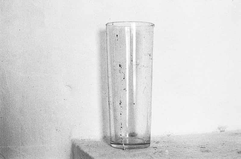 Ein schlankes Wasserglas steht auf einem Mauersims vor einer Wand. Sims und Wand sind gekaelkt. Das Glas ist mit Staub und Dreck bedeckt. Einfallendes Licht bringt Zeichnung in das Glas und wirft einen schoenen Schatten auf die Wand. Es handelt sich um eine Schwarz - Weiss Fotografie.