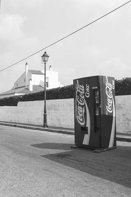 Ein Coca Cola Automat steht vor einer weissen Mauer, die von einer Hecke ueberragt wird auf der Strasse. Links von ihm befindet sich eine Strassenlaterne und hinter der Mauer befindet sich ein Haus, wie es auf Menorca gebaut wird. Im oberen Bildbereich ist eine elektrische Ueberlandleitung mit diagonalem Verlauf zu sehen. Es handelt sich um eine Schwarz - Weiss Fotografie.