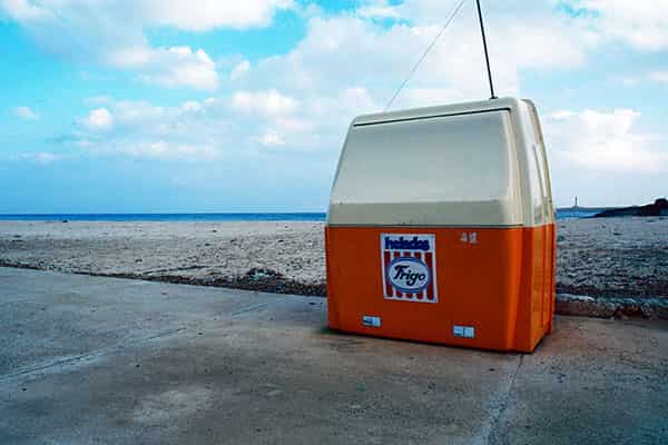 Eine verlassene Strandsituation auf Menorca im Winter. Der Bildaufbau ist wie folgt: im Vordergrund befindet sich eine betonierte Strasse - ihr folgt ein breiter Streifen Sandstrand. Dahinter ist ein schmaler blauer Streifen Mittelmeer zu sehen, über ihm bewoelkter Himmel. Die Szene ist horizontal ausgerichtet. Auf der Strasse, an der Grenze zum Strand, steht ein Verkaufsstand - einer Box gleich - aus Plastik fuer den Verkauf von Eis der Marke Frigo. Die untere Haelfte ist orange, die obere Haelfte weiss und laesst sich nach vorne aufklappen, so das eine Art Tresen entsteht. Auf dem Dach der Box ist eine Antenne installiert.