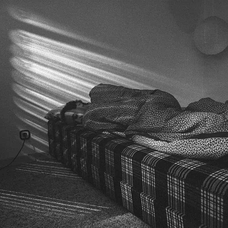 Ein Mann liegt auf einer doppelten Matratze. Sie ist mehrfach kariert und liniert. Eine Bettdecke liegt über seinem gesamten Koerper - nur der Kopf ist zu sehen. Die Bettdecke ist gebluemt. Licht faellt in Form von markanten Strahlen in das Zimmer. Im oberen rechten Eck befindet sich eine Papierlampe, links neben dem Bett eine Steckdose. Es handelt sich um eine Schwarz - Weiss Fotografie.