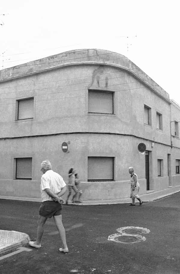 Eine unbefahrene Strassenkreuzung auf Menorca, in der Mitte befinden sich zwei nebeneinander liegende Gullydeckel. Ein zweigeschossiges Haus ohne Ecke praegt dominant das S/W-Foto. Die Ecke des Hauses ist gerundet, bei allen Fenstern sind die Rolladen geschlossen. Eine dicke Stromleitung verlaeuft waagerecht zwischen Parterre und dem erstem Stock an der Aussenwand entlang - Verbotsschilder sind fuer zwei der Abzweigungen an ihr angebracht. Links im Vordergrund ist ein aelterer Herr, der seine Arme hinter dem Ruecken verschraenkt hat, gerade dabei, den schmalen Buergersteig zu betreten. Er trägt eine dunkle kurze Hose und ein weisses kurzaermeliges Hemd. Auf der gegenüberliegenden Strassenseite laufen zwei Kinder in die entgegengesetzte Richtung. Ihre Abbildung ist wegen ihrer schnelleren Bewegung unscharf. Ihnen entgegen kommt eine ältere Dame, mit einer leicht gebeugten Haltung. Sie trägt ein luftiges, wenig gemustertes, kurzaermeliges weisses Kleid.