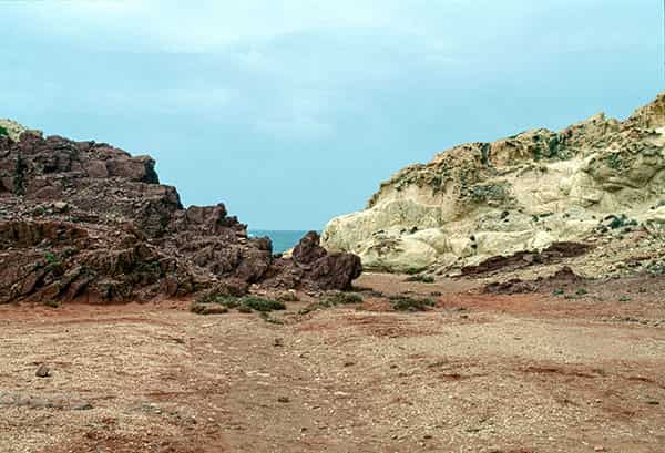 Eine unbefahrene Strassenkreuzung auf Menorca, in der Mitte befinden sich zwei nebeneinander liegende Gullydeckel. Ein zweigeschossiges Haus ohne Ecke praegt dominant das S/W-Foto. Die Ecke des Hauses ist gerundet, bei allen Fenstern sind die Rolladen geschlossen. Eine dicke Stromleitung verlaeuft waagerecht zwischen Parterre und dem erstem Stock an der Aussenwand entlang - Verbotsschilder sind fuer zwei der Abzweigungen an ihr angebracht. Links im Vordergrund ist ein aelterer Herr, der seine Arme hinter dem Ruecken verschraenkt hat, gerade dabei, den schmalen Buergersteig zu betreten. Er trägt eine dunkle kurze Hose und ein weisses kurzaermeliges Hemd. Auf der gegenüberliegenden Strassenseite laufen zwei Kinder in die entgegengesetzte Richtung. Ihre Abbildung ist wegen ihrer schnelleren Bewegung unscharf. Ihnen entgegen kommt eine ältere Dame, mit einer leicht gebeugten Haltung. Sie trägt ein luftiges, wenig gemustertes, kurzaermeliges weisses Kleid.