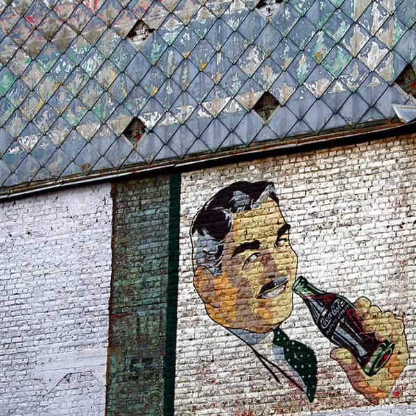 Eine unbefahrene Strassenkreuzung auf Menorca, in der Mitte befinden sich zwei nebeneinander liegende Gullydeckel. Ein zweigeschossiges Haus ohne Ecke praegt dominant das S/W-Foto. Die Ecke des Hauses ist gerundet, bei allen Fenstern sind die Rolladen geschlossen. Eine dicke Stromleitung verlaeuft waagerecht zwischen Parterre und dem erstem Stock an der Aussenwand entlang - Verbotsschilder sind fuer zwei der Abzweigungen an ihr angebracht. Links im Vordergrund ist ein aelterer Herr, der seine Arme hinter dem Ruecken verschraenkt hat, gerade dabei, den schmalen Buergersteig zu betreten. Er trägt eine dunkle kurze Hose und ein weisses kurzaermeliges Hemd. Auf der gegenüberliegenden Strassenseite laufen zwei Kinder in die entgegengesetzte Richtung. Ihre Abbildung ist wegen ihrer schnelleren Bewegung unscharf. Ihnen entgegen kommt eine ältere Dame, mit einer leicht gebeugten Haltung. Sie trägt ein luftiges, wenig gemustertes, kurzaermeliges weisses Kleid.