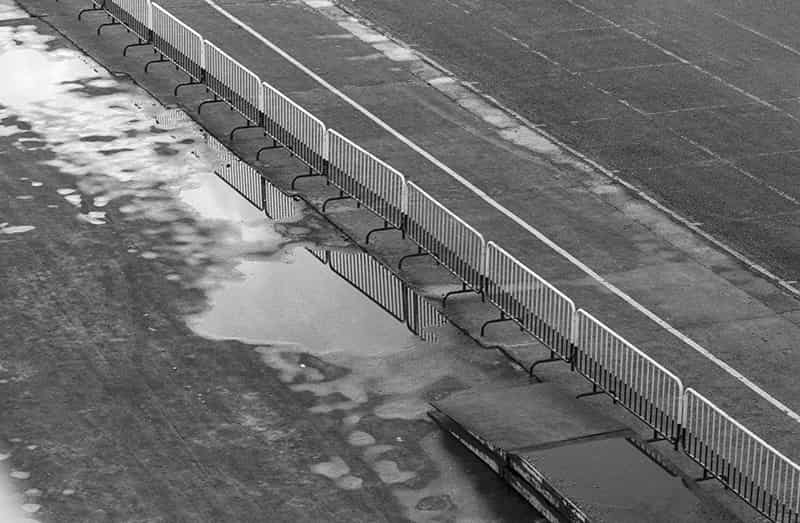 Eine Flaeche aus Asphalt ist durch Absperrgitter diagonal zweigeteilt. In der rechten Haelfte der Schwarzweiss Fotografie befindet sich so etwas wie eine Fahrbahn mit einer weissen durchgezogenen Linie in der Mitte. Eine zweite Linie begrenzt einen Parkplatz mit der entsprechenden weissen Unterteilung fuer die Fahrzeuge.  In der linken Haelfte befindet sich eine Flaeche aus Asphalt mit Pfuetzen, in denen sich die Absperrgitter spiegeln. Im rechten unteren Bildbereich sind zwei uebereinander gestapelte Rampen.