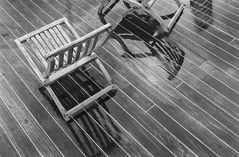 Zwei Deckchairs stehen auf nassem Schiffsparkett - einer ist angeschnitten. Sie spiegeln sich im nassen Untergrund. Es handelt sich um eine Schwarz - Weiss Fotografie.