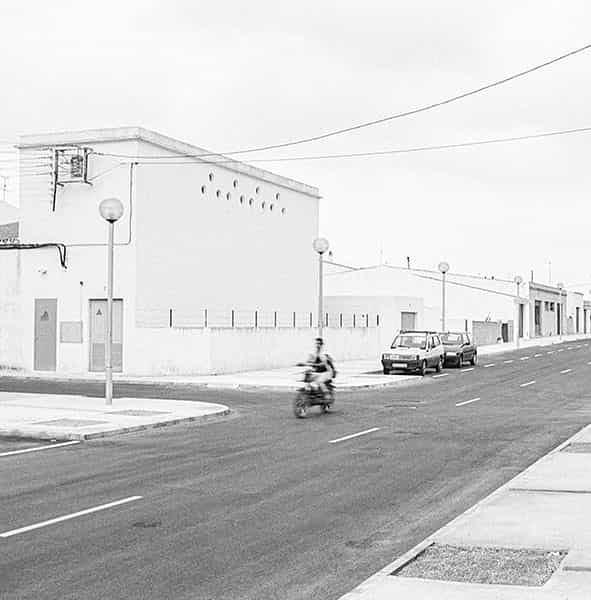 Eine unbefahrene Strassenkreuzung auf Menorca, in der Mitte befinden sich zwei nebeneinander liegende Gullydeckel. Ein zweigeschossiges Haus ohne Ecke praegt dominant das S/W-Foto. Die Ecke des Hauses ist gerundet, bei allen Fenstern sind die Rolladen geschlossen. Eine dicke Stromleitung verlaeuft waagerecht zwischen Parterre und dem erstem Stock an der Aussenwand entlang - Verbotsschilder sind fuer zwei der Abzweigungen an ihr angebracht. Links im Vordergrund ist ein aelterer Herr, der seine Arme hinter dem Ruecken verschraenkt hat, gerade dabei, den schmalen Buergersteig zu betreten. Er trägt eine dunkle kurze Hose und ein weisses kurzaermeliges Hemd. Auf der gegenüberliegenden Strassenseite laufen zwei Kinder in die entgegengesetzte Richtung. Ihre Abbildung ist wegen ihrer schnelleren Bewegung unscharf. Ihnen entgegen kommt eine ältere Dame, mit einer leicht gebeugten Haltung. Sie trägt ein luftiges, wenig gemustertes, kurzaermeliges weisses Kleid.