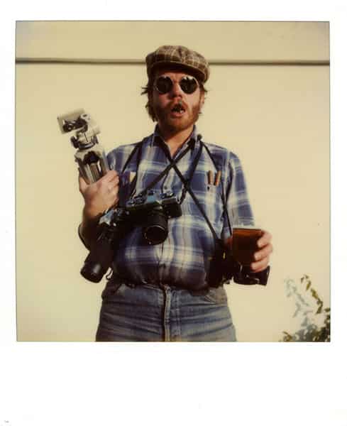 Der Reporter - drei Kameras haengen um seinen Hals, das Stativ ist unter den Arm geklemmt. In seiner linken Hand ein Glas Bier, zwei Zigarren aus Cuba in seiner Hemdtasche, eine Zigarre im Mund, der von einem roten Bart eingerahmt ist. Die Augen sind von einer runden Sonnenbrille verdeckt, sein Haupt ziert eine braune Kappe, die mit zebraaehnlichen Streifen versehen ist. Der Reporter traegt ein blau - weiss kariertes Hemd und eine Blue Jeans. Der Bauch wird durch ein Kissen unter dem Hemd zur Grösse. Das Selbstportrait wurde vor einer Garagenwand auf Menorca mittels einer Polaroidkamera aufgenommen.
