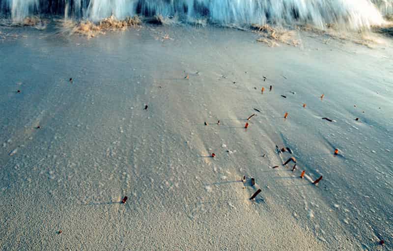 Das obere sechstel der Farbfotografie zeigt eine hereinbrechende kleine Welle auf einen flachen Sandstrand. Sie trifft von oben einfallend auf den Sand, der aufspritzt. Die Welle und der aufgewuehlte Sand sind ob ihrer Dynamik unscharf. Das vordere Sechstel ist Sandstrand, Wasser, welches durch die zuvordere Welle weiter auf den Strand ausduente stroemt zurueck, welches wiederum durch Unschaerfe gekennzeichnet ist. Die Sonne steht tief und beleuchtet die Szene von rechts. Flaches Seegras, welches im Sand feststeckt wird teilweise durchleuchted und wirft lange Schatten.
