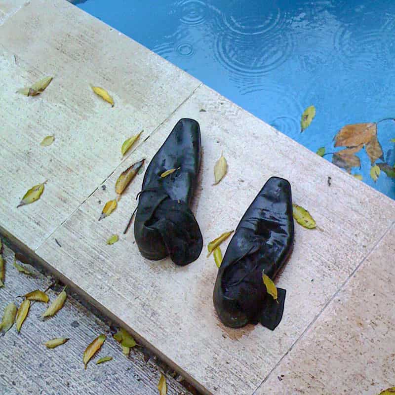 Im Zentrum der Farbfotografie befindet sich diagonal eine Flaeche aus Travertin, auf der ein Paar schwarze Schuhe steht, welche durchnaesst sind und aus dem schwarze nasse Socken haengen. Im rechten oberen Eck ist Wasser eines Pool. Es regnet und die Tropfen bringen Kreise auf der Wasseroberflaeche hervor. Im linken unteren Dreieck befindet sich eine Steinplatte. In allen drei Ebenen bedecken herab gefallene Blaetter das Wasser, die Schuhe, die Struempfe, den Travertin