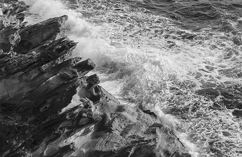 Felsen, Meer, Brandung und Gischt praegen das Foto. Das dunkle Gestein ragt gezackt diagonal in das Foto. An ihm zerbrechen die Wellen mit weissem Schaum, der langsam abfliesst und in sanften Wellen abebbt. Es handelt sich um eine Schwarz - Weiss Fotografie.