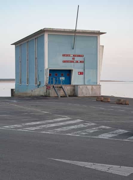 Eine unbefahrene Strassenkreuzung auf Menorca, in der Mitte befinden sich zwei nebeneinander liegende Gullydeckel. Ein zweigeschossiges Haus ohne Ecke praegt dominant das S/W-Foto. Die Ecke des Hauses ist gerundet, bei allen Fenstern sind die Rolladen geschlossen. Eine dicke Stromleitung verlaeuft waagerecht zwischen Parterre und dem erstem Stock an der Aussenwand entlang - Verbotsschilder sind fuer zwei der Abzweigungen an ihr angebracht. Links im Vordergrund ist ein aelterer Herr, der seine Arme hinter dem Ruecken verschraenkt hat, gerade dabei, den schmalen Buergersteig zu betreten. Er trägt eine dunkle kurze Hose und ein weisses kurzaermeliges Hemd. Auf der gegenüberliegenden Strassenseite laufen zwei Kinder in die entgegengesetzte Richtung. Ihre Abbildung ist wegen ihrer schnelleren Bewegung unscharf. Ihnen entgegen kommt eine ältere Dame, mit einer leicht gebeugten Haltung. Sie trägt ein luftiges, wenig gemustertes, kurzaermeliges weisses Kleid.