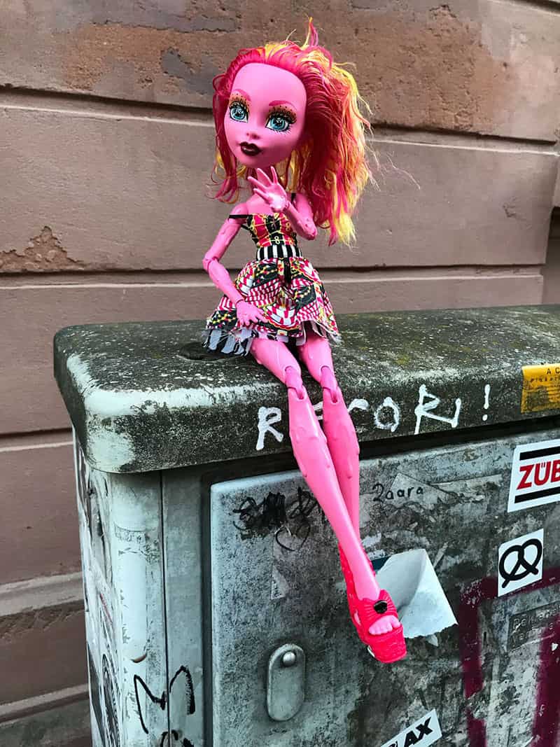 Eine Puppe mit unnatuerlicher Hautfarbe sitzt auf einem dreckigen Verteilerkasten vor einer braunen Wand. Sie hat sehr lange Beine und ihre Fuesse sind verschrenkt. Sie traegt rote Sandalen. Sie hat grosse blaue Augen und ihr Haar ist rot und gelb gefaerbt. 