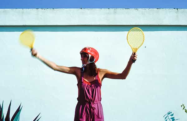 Eine sportliche schlanke Frau, gekleidet in einer lila Latzhose, steht vor einer weissen Wand. Sie hält zwei gelbe Kindertennisschläger in ihren Haenden. Ihre muskuloesen Arme hat sie weit vom Koerper gestreckt, die Plastiktennisschlaeger weisen nach oben, als ob sie ein imaginaeres Flugzeug einweisen will. Die Spitzen einer Agave ragen unten links in das Bild, welches auf Menorca aufgenommen wurde