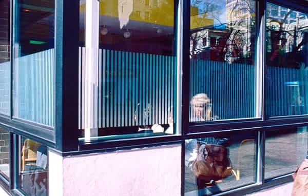 New York in den 80er Jahren. Eine Straßenecke – Art Deko – klassisch, die Fenster geben einen verspiegelten Einblick in ein New Yorker Cafe. Ein blondierter Frauenkopf ist zu sehen – sitzt sie im Cafe oder geht sie an ihm vorbei, es ist nicht direkt ersichtlich. Aber es ist genau dies, der unklare Uebergang zwischen Innen und Aussen, der dieses Foto besonders macht und den Charakter New Yorks in sich spiegelt. Spieglein Spieglein an der Wand, wer ist die Schoenste im ganzen Land? New York!