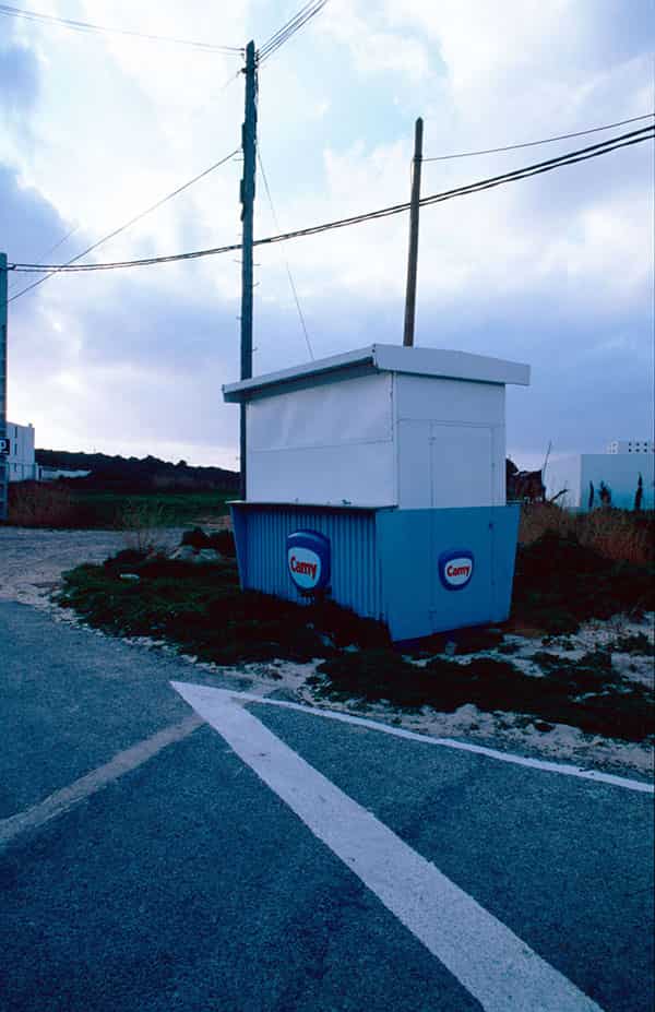 Eine Strasse auf der winterlichen Insel Menorca - sie bildet eine lang gestreckte Kurve und eine weisse Strassenmarkierung in Form eines Pfeils ist dominant im Vordergrund dieser Farbfotografie zu sehen. In der Kurve steht ein Verkaufsstand. Der Stand ist wie eine Box, unten blau, oben weiss. Ein Logo ist im unteren Bereich an der Front und der Seite angebracht. Ein weisses Feld ist dunkelblau umrandet und der rote Schriftzug „Camy“ steht in der Mitte. Der obere Bereich des Fotos besteht zu 50 % aus bewölktem Himmel. In ihn herein ragen hoelzerne Strommasten, Ueberlandkabel durchschneiden ihn.
