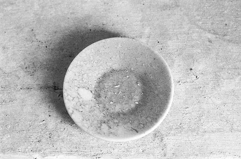 Auf einem weissen gekaelkten Untergrund steht ein weisser Unterteller. Er ist mit Staub bedeckt. Einfallendes Licht wirft einen Schatten auf den Untergrund. Es handelt sich um eine Schwarz - Weiss Fotografie.