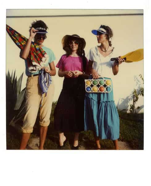 Drei Grazien vor der Garagenwand auf Menorca, gebannt auf Polaroid. Von links beginnend: der rechte Arm der grossen schlanken Lady ist angewinkelt und traegt in der Armbeuge einen bunt gemusterten Sonnenschirm. Die gelbe Hose ist unten aufgekrempelt, die Beine sind braun, so wie ihre nackten Arme. Sie traegt ein tuerkisfarbenes T-Shirt. Die Sonnenbrille rutscht ihr von der Nase, der Blick geht über ihr hinaus. Ihre rechte Hand greift an die Kappe ihres Sonnenschutzes. Die Lady neben ihr traegt einen Strohhut und ihre Augen sind durch eine runde Sonnenbrille verdeckt. Sie traegt ein lilanes t-shirt und einen dunklen langen Rock, ihre Haende sind unterhalb ihrer Brueste verschraenkt. Sie ist die kleinste im Bunde. Die Frau an ihrer rechten Seite schaut in Richtung der ersten Person. Sie traegt auch eine Sonnenkappe, das Visier in blau. In ihrer rechten Hand hält sie ein Netz mit Bocciakugeln - eine Kugel fehlt und der blaue Stoff ihres Kleides ersetzt die Farbe der fehlenden Kugel. In der rechten Hand haelt sie einen Soft-ball-Schlaeger, der auf Grund der Bewegung ihres Armes unscharf ist. Der Oberkoerper wird von einem weissen Top mit V-Ausschnitt bedeckt.