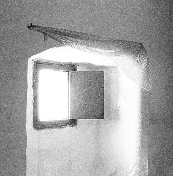 Das Schlafzimmer in einer verlassenen Finca auf Menorca. Nur ein Wandausschnitt ist auf diesem S/W-Foto abgebildet. Ein Fenster befindet sich in einer Art von Erker - zurueckgesetzt. Durch eine Klappe aus Holz laesst es sich verschliessen. Die Klappe steht im 135 Grad Winkel offen und laesst gleissendes Licht in den Erker stroemen. Nur der Erker erstrahlt in diesem Licht, die Waende, welche ihn einfassen bleiben in Grau. Ein Aussen kann man nicht mehr erkennen, so hell ist das einströmende Licht. Der Fensterrahmen wirkt wie ausgefranst und das dunkle Holz der Klappe ist stark aufgehellt. Oberhalb der Nische ist eine gehaekelte Gardine an Metallhaken angebracht. Sie faellt nicht senkrecht herunter, sondern hängt zusammen gerafft ueber dem rechten Haken. Durch das einfallende Licht erscheint ihre Struktur von Dunkelgrau ueber Hellgrau bis zu Weiss mit wenig Struktur.