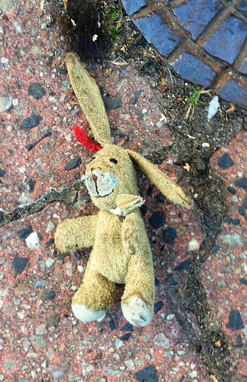 Ein Spielzeug Hase aus gelbem Stoff liegt wie achtlos weggeworfen auf dem Gehsteig. Ein Ohr steht fast senkrecht nach oben, das andere zeigt in einem Winkel von 45 Grad nach unten. 