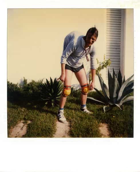 Quasimodo - sie gab mir Wasser. Da steht er vor der Garagenwand mit seinem Buckel, gebeugt. Er traegt Knieschuetzer, kurze schwarze Hose, graublaues t-shirt und Turnschuhe. Seine Haltung ist gebeugt. Die Polaroidaufnahme zeigt ihn vor einer Garagenwand auf Menorca. Links und rechts befinden sich Agaven. Im rechten Teil der Wand befindet sich ein vertikaler Einschnitt, der mit hoelzernen, waagerechten Lamellen fuer Luftaustausch sorgen soll.